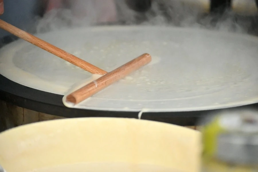 Préparation d’une crêpe de froment sur un billig chaud, avec étalement de la pâte à l’aide d’un rozell en bois, pendant la cuisson.