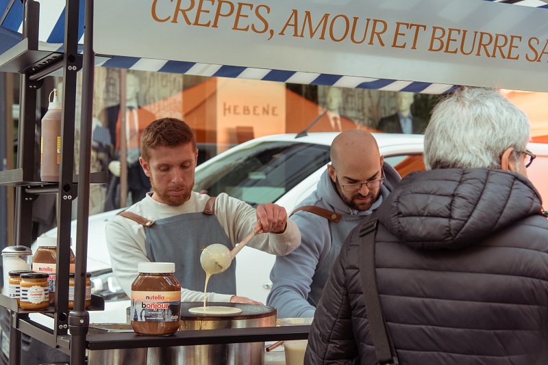 Deux crêpiers en tabliers préparent une crêpe artisanale sur une billig traditionnelle lors d'un marché en Bretagne. Au premier plan, des pots de Nutella et de caramel au beurre salé. Enseigne "Crêpes, Amour et Beurre Salé" visible en arrière-plan.